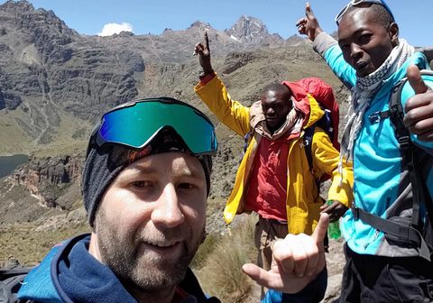 Michal in a selfie on Mount Kenya, with ski goggles on his beanie, two local guides cheering behind him with a thumbs up and rocky peaks in the distance