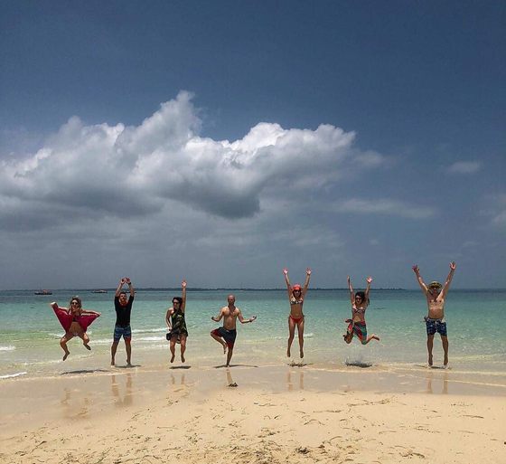 Seven friends, Michal among them, jumping in the air on a sandy beach in Zanzibar, with turquoise water and a big cloud behind