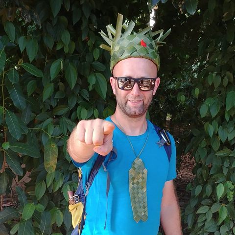 Michal wearing a woven palm-leaf crown and a palm-leaf tie, pointing straight at the viewer with a grin, in front of dense jungle leaves